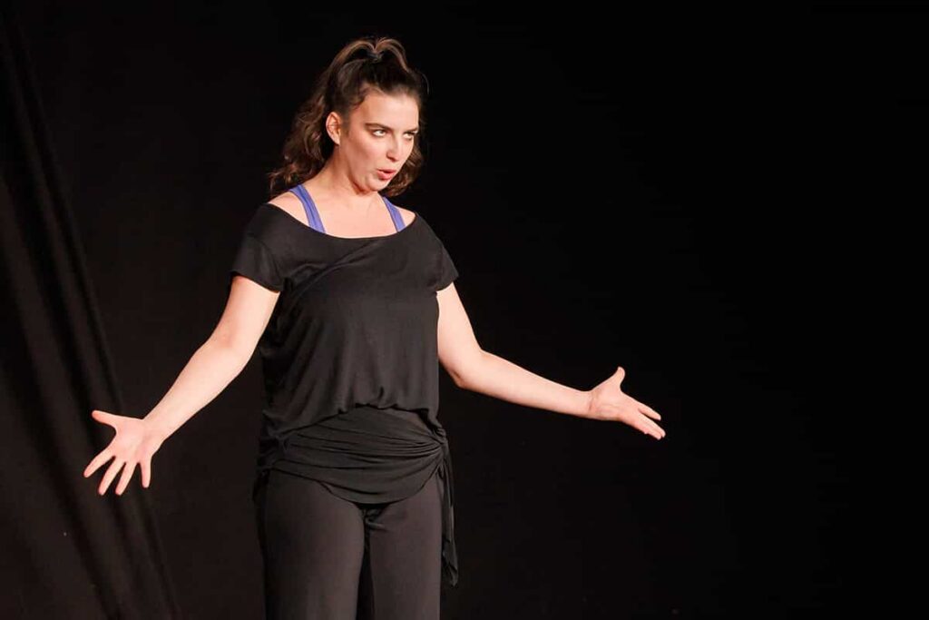 Alexia Mocănescu pe scenă, îmbrăcată în negru, cu brațele întinse și o expresie teatrală, în fața unui fundal întunecat.