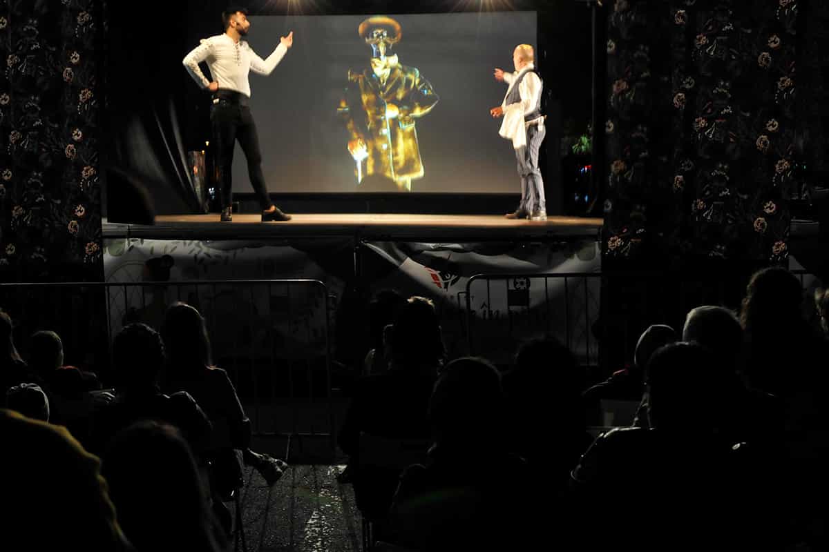 Doi actori pe scenă, în fața unei proiecții, în timpul unui spectacol urmărit de public la Bucharest Fringe Festival.